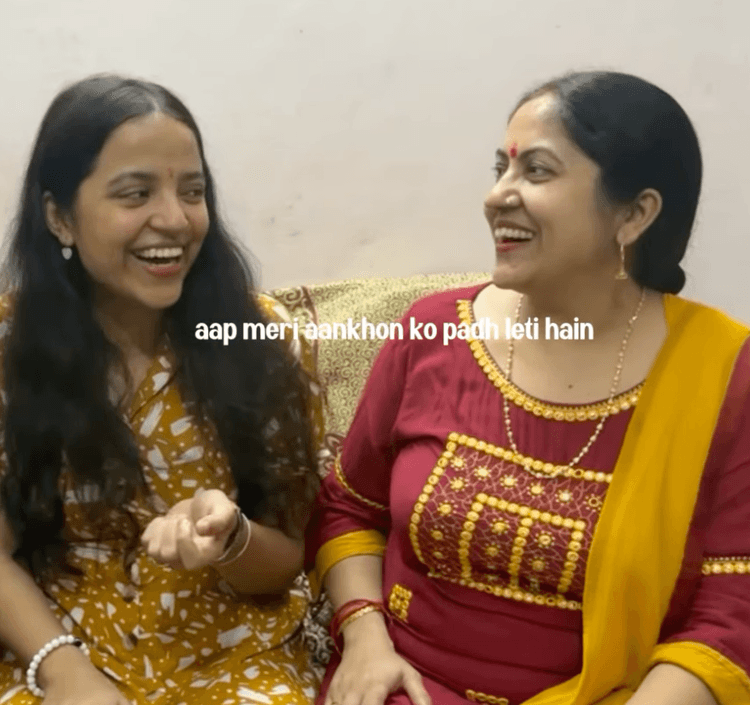 Shivangi in a mustard floral dress and her mother in a maroon kurta, laughing together on the sofa